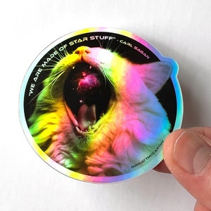 May include: A holographic sticker featuring a cat with its mouth open, revealing a galaxy inside. The sticker has the text "We are made of star stuff." Carl Sagan around the cat's head.