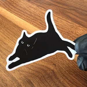 Op de afbeelding: Een zwarte kat sticker met witte contouren, de kat ligt op zijn zij met zijn hoofd naar links gedraaid, de kat heeft witte ogen en een witte mond.