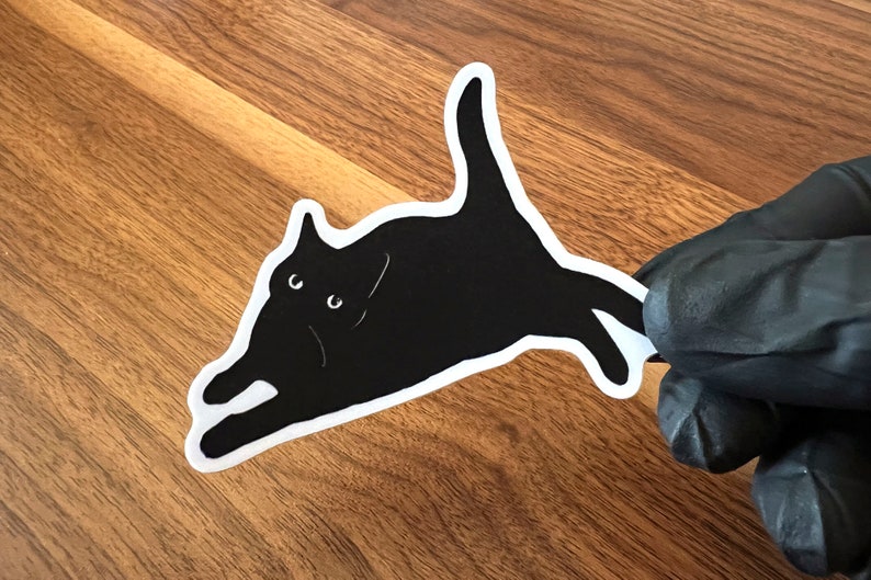 Op de afbeelding: Een zwarte kat sticker met witte contouren, de kat ligt op zijn zij met zijn hoofd naar links gedraaid, de kat heeft witte ogen en een witte mond.