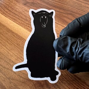 Op de afbeelding: Zwart-witte silhouetsticker van een kat met zijn bek open, alsof hij gaapt. De sticker zit op een houten oppervlak.