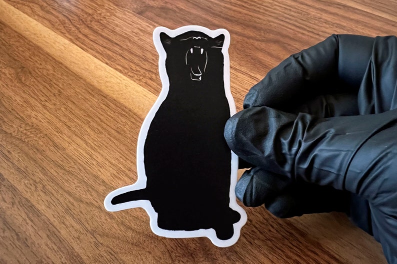Op de afbeelding: Zwart-witte silhouetsticker van een kat met zijn bek open, alsof hij gaapt. De sticker zit op een houten oppervlak.