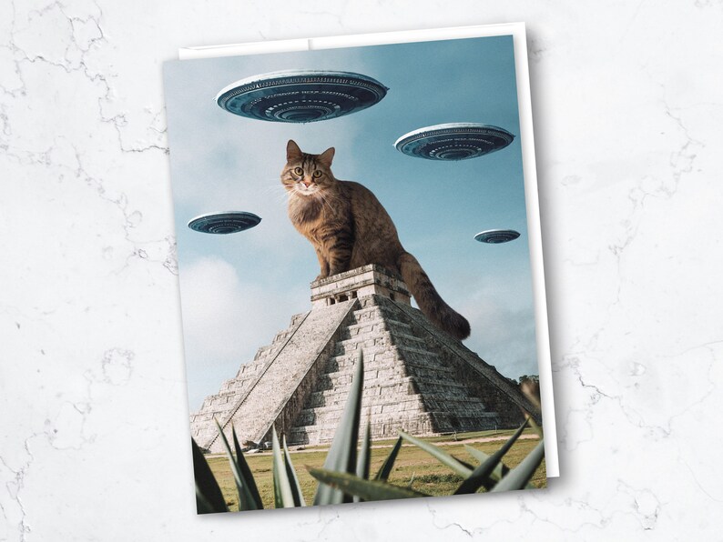May include: A brown tabby cat sits atop a stone pyramid, with several flying saucers hovering in the sky above. The pyramid is surrounded by green plants.