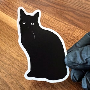 Op de afbeelding: Zwarte kat sticker met een witte omtrek. De kat zit met zijn oren omhoog en kijkt recht naar de kijker.