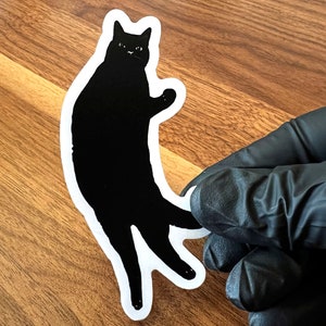 Op de afbeelding: Een zwarte kat sticker met een witte omtrek. De kat staat op zijn achterpoten met zijn voorpoten omhoog.