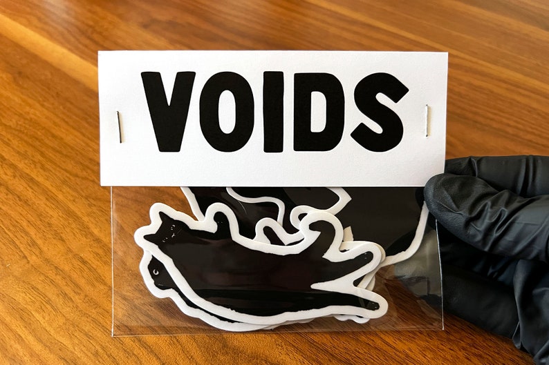 Op de afbeelding: Een pakket met zwarte en witte katten stickers met het woord "VOIDS" gedrukt op de voorkant.