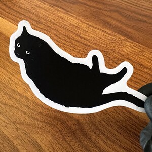 Op de afbeelding: Een zwarte kat sticker met witte contouren, de kat ligt op zijn rug met zijn poten in de lucht.