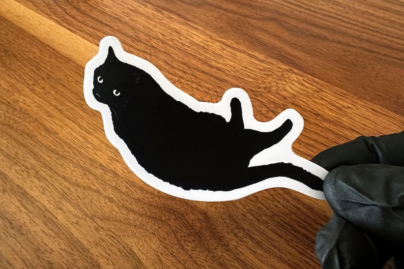 Op de afbeelding: Een zwarte kat sticker met witte contouren, de kat ligt op zijn rug met zijn poten in de lucht.