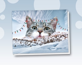 May include: A Christmas card featuring a large cat's head peeking over a snowy mountain range. The cat has gray and white fur and green eyes. A string of colorful Christmas lights is strung across the top of the image.