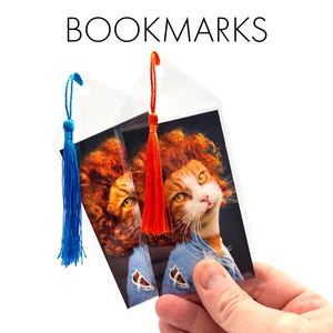 May include: Two bookmarks with a cat wearing a lion's mane wig. The cat is orange and white, with a denim jacket. The bookmarks have blue and orange tassels and the word BOOKMARKS is at the top.