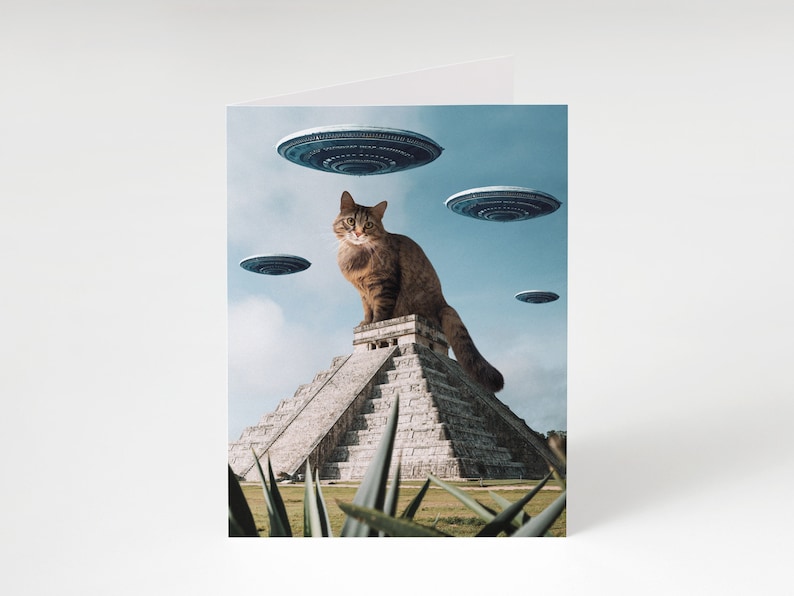 May include: A brown tabby cat sits atop a stone pyramid, looking directly at the viewer. Several flying saucers hover in the sky above the cat.