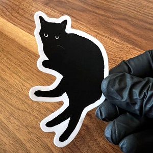 Op de afbeelding: Zwarte kat sticker met een witte omtrek. De kat zit met zijn poten onder zijn lichaam getrokken en zijn hoofd gekanteld naar de zijkant. De sticker zit op een houten oppervlak.