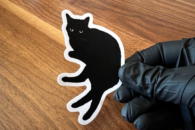 Op de afbeelding: Zwarte kat sticker met een witte omtrek. De kat zit met zijn poten onder zijn lichaam getrokken en zijn hoofd gekanteld naar de zijkant. De sticker zit op een houten oppervlak.