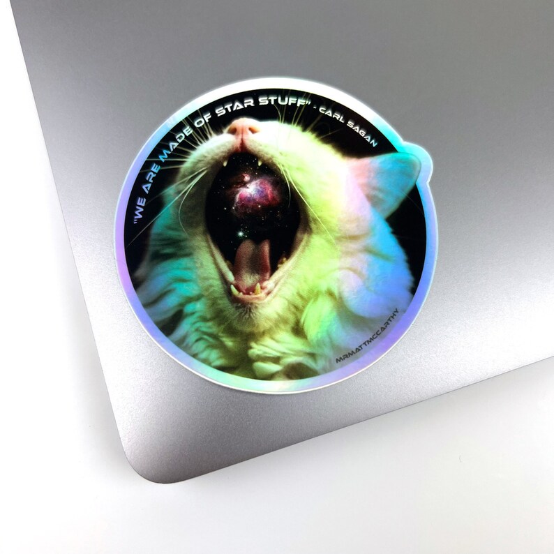May include: A holographic sticker featuring a white cat with its mouth open, revealing a galaxy inside. The sticker has the text "We are made of star stuff" and "Carl Sagan" around the cat's head.