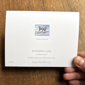 May include: A Christmas card featuring a cat peeking out from a snowy landscape with the text "Happy Holidays". The card is designed by MrMattMcCarthy, a surreal cat art creator. The card is designed and printed in the USA.