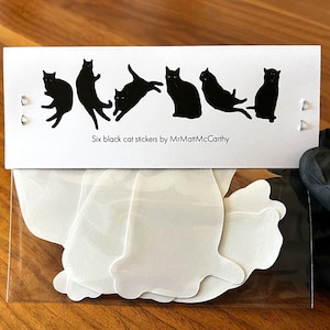 Op de afbeelding: Een doorzichtige plastic zak met zes witte katten stickers. De zak ligt op een houten oppervlak. De stickers zijn in de vorm van zwarte katten in verschillende poses. Achter de zak zit een kaart met de tekst "Six black cat stickers by MrMattMcCarthy".