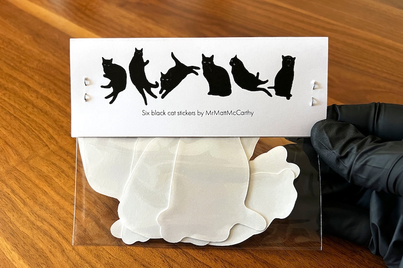 Op de afbeelding: Een doorzichtige plastic zak met zes witte katten stickers. De zak ligt op een houten oppervlak. De stickers zijn in de vorm van zwarte katten in verschillende poses. Achter de zak zit een kaart met de tekst "Six black cat stickers by MrMattMcCarthy".