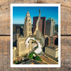 May include: A tabby cat with white markings is positioned in a yoga pose, seemingly holding up a tall building in a city skyline. The cat's head is tilted back, and its eyes are closed. The building is a tall, slender structure with a pointed roof and many windows. The city skyline is visible in the background, with other buildings and a blue sky.