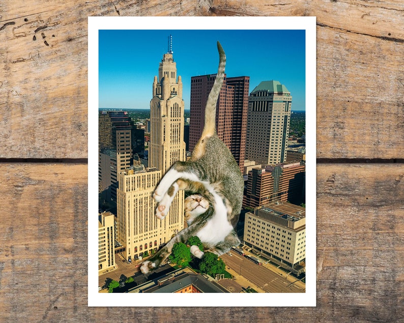 May include: A tabby cat with white markings is positioned in a yoga pose, seemingly holding up a tall building in a city skyline. The cat's head is tilted back, and its eyes are closed. The building is a tall, slender structure with a pointed roof and many windows. The city skyline is visible in the background, with other buildings and a blue sky.