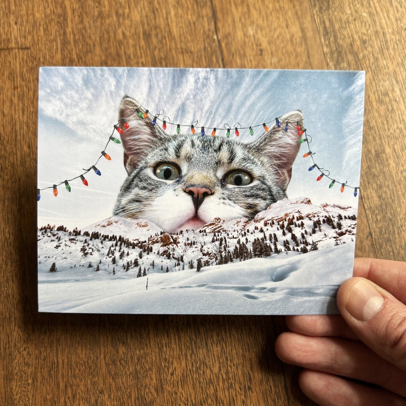 May include: A humorous Christmas card featuring a large cat's face peeking over a snowy mountain landscape. The cat has a surprised expression and is wearing a string of colorful Christmas lights.