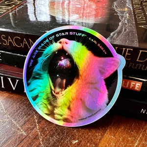 May include: A rainbow holographic sticker featuring a cat's face with an open mouth. The cat's mouth is filled with a galaxy. The sticker includes the text "We are made of star stuff" and "Carl Sagan" around the cat's face.