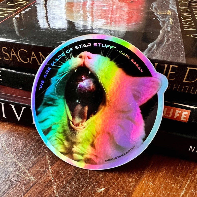 May include: A rainbow holographic sticker featuring a cat's face with an open mouth. The cat's mouth is filled with a galaxy. The sticker includes the text "We are made of star stuff" and "Carl Sagan" around the cat's face.