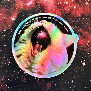 May include: A holographic sticker featuring a white cat yawning with a galaxy inside its mouth. The sticker is round and has the text "We are made of star stuff" Carl Sagan" around the edge.