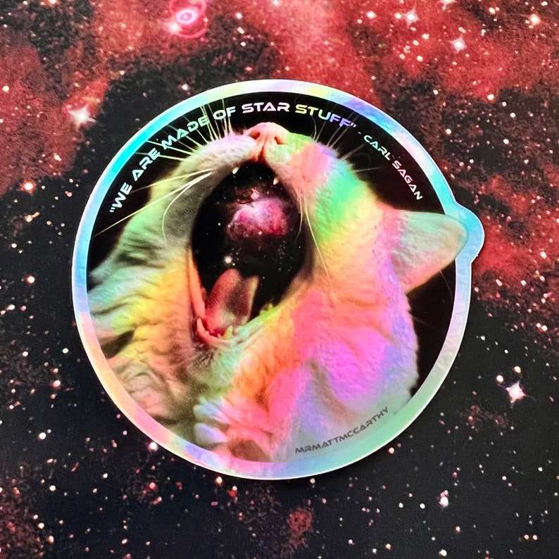 May include: A holographic sticker featuring a white cat yawning with a galaxy inside its mouth. The sticker is round and has the text "We are made of star stuff" Carl Sagan" around the edge.