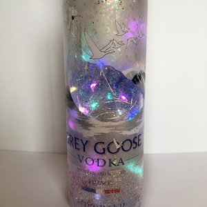Puede incluir: Una botella de vidrio transparente con una etiqueta plateada que dice "Grey Goose Vodka" y "Destilado y embotellado en Francia". La botella está decorada con purpurina y detalles iridiscentes, y presenta un diseño de gansos volando y una cordillera.