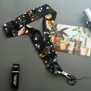 Pode incluir: Lanyard preto e dourado com estampa de galáxia com estrelas e planetas. O lanyard tem um fecho giratório prateado e um cordão preto.