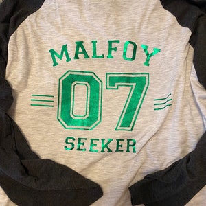 May include: A gray and black baseball jersey with green text that reads "Malfoy 07 Seeker".