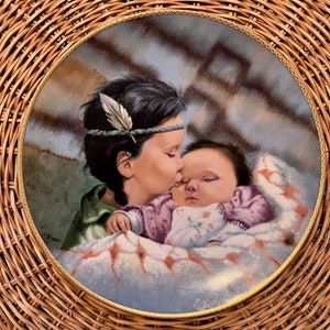 Peut inclure: Une assiette en porcelaine avec un bord doré présente une peinture d'une jeune fille avec un bandeau à plumes embrassant un bébé endormi enveloppé dans une couverture. L'arrière-plan est un tissu à motifs bleus et bruns.