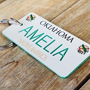 May include: A white keychain with the name "AMELIA" in teal, and the text "OKLAHOMA" and "NATIVE AMERICA" in black and gold. The keychain has a silver ring and a Native American design.