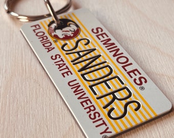 May include: A white metal keychain with a yellow and red striped design. The keychain features the text "Seminoles" and "Sanders" in black lettering. The text "Florida State University" is also included in red lettering.