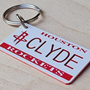 May include: White Houston Rockets keyring with red lettering that reads "HOUSTON CLYDE ROCKETS". The keyring features a red Houston Rockets logo.