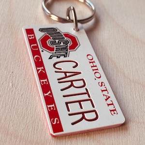 Puede incluir: Llavero blanco de los Ohio State Buckeyes con un logotipo de Ohio State rojo y blanco y el nombre "Carter" en letras rojas.