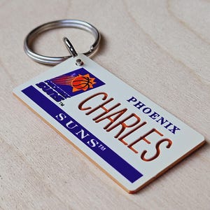 Könnte beinhalten: Ein weißer und lilafarbener Schlüsselanhänger mit einem Basketball-Logo und dem Text "Phoenix Suns" und "Charles".