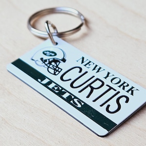 May include: White and green New York Jets keychain with a silver key ring. The keychain features a New York Jets logo and the name "Curtis" on a white background.