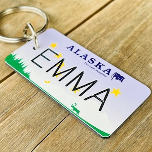 Peut inclure: Un porte-clés blanc et bleu avec le texte "ALASKA The Last Frontier" et le nom "EMMA" en noir. Le porte-clés a un fond vert et blanc avec une petite image d'un élan.