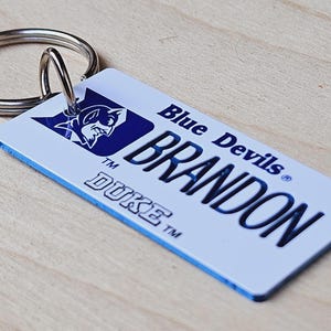 May include: A white and blue Duke Blue Devils keychain with the name "BRANDON" printed in blue. The keychain features a blue devil mascot logo and the text "Blue Devils" and "DUKE" in blue.