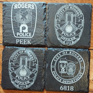 May include: Four black slate coasters with white engraved designs. The coasters feature the City of Rogers Police Department logo, including the department's motto, "Police Officer", and the city's founding year, 1914. The coasters also include the department's badge number, 6818.