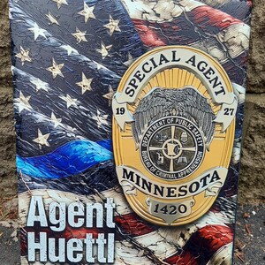 May include: A decorative plaque featuring an American flag design with a blue line. A gold-colored badge with the text "SPECIAL AGENT" and "MINNESOTA" is affixed to the flag. The name "Agent Huettl" is displayed.