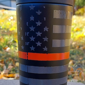 May include: A black insulated tumbler with a thin red stripe and a faded American flag design. The tumbler has the text "FrostBuddy." printed on the bottom.