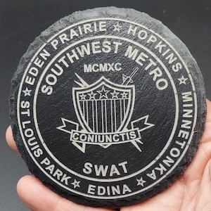 May include: Black slate coaster with a white engraved design. The design features a shield with five stars, two crossed swords, and the text "Southwest Metro", "MCMXC", "Coniunctis", "SWAT", "Eden Prairie", "Hopkins", "St. Louis Park", "Edina", and "Minnetonka".