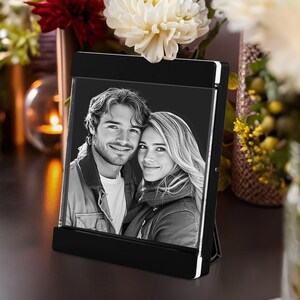 May include: A black and white photo frame with a picture of a smiling couple. The frame is rectangular with a black base and a white border. The photo is displayed vertically and is surrounded by flowers.