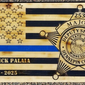May include: A wooden American flag with a blue line, a sheriff's star badge, and the text "253 MAJOR DEPUTY SHERIFF STATE OF MINNESOTA HENNEPIN COUNTY" and "MAJOR RICK PALAIA 1998 - 2025".