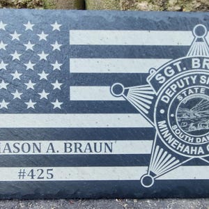 May include: A rectangular black slate plaque featuring an engraved American flag design. The text SGT. MASON A. BRAUN and #425 are etched below the stars. A Deputy Sheriff star badge for Minnehaha County, South Dakota, is engraved on the right.