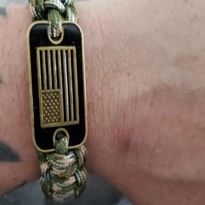 May include: Camouflage paracord bracelet with a black rectangular tag featuring a gold-colored American flag design. The bracelet is a mix of green, brown, and tan colors.