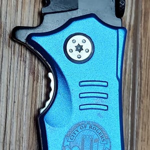 May include: A blue and black pocket knife with a serrated blade and a bottle opener. The knife has a silver emblem with the words "City of Rogers Police Officer 20" and the text "PEEK 20" on the blade.