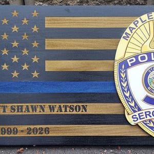 May include: Wooden American flag with gold stars, blue stripe, and black and gold stripes. Features a Maple Grove Police badge and text: Sergeant Shawn Watson 1999-2020.