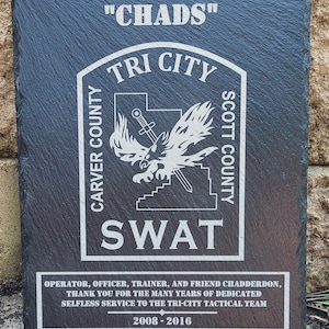 May include: A black slate plaque with a white engraved inscription. The inscription reads "CARVER COUNTY" "CHADS" "TRI CITY" "SWAT" "SCOTT COUNTY" "OPERATOR, OFFICER, TRAINER, AND FRIEND CHADDERDON, THANK YOU FOR THE MANY YEARS OF DEDICATED SELFLESS SERVICE TO THE TRI-CITY TACTICAL TEAM 2008-2016".